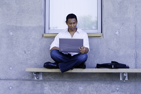 A man sits outside of a campus building with a laptop.