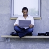 A man sits outside of a campus building with a laptop.